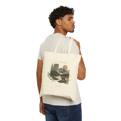 Natural cotton canvas tote bag printed with a layered Alaska mountain and pine forest scene by Evernorth — styled flat lay